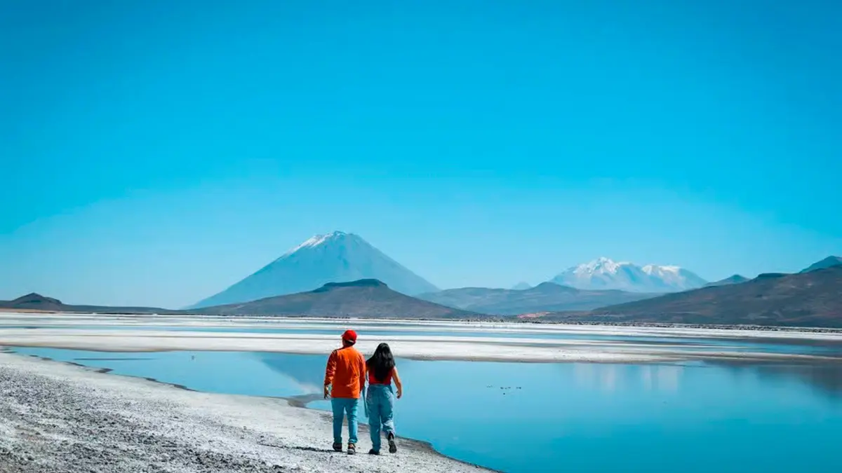 Pareja de excursionistas caminando tomados de la mano por el borde de la Laguna de Salinas y las montañas.