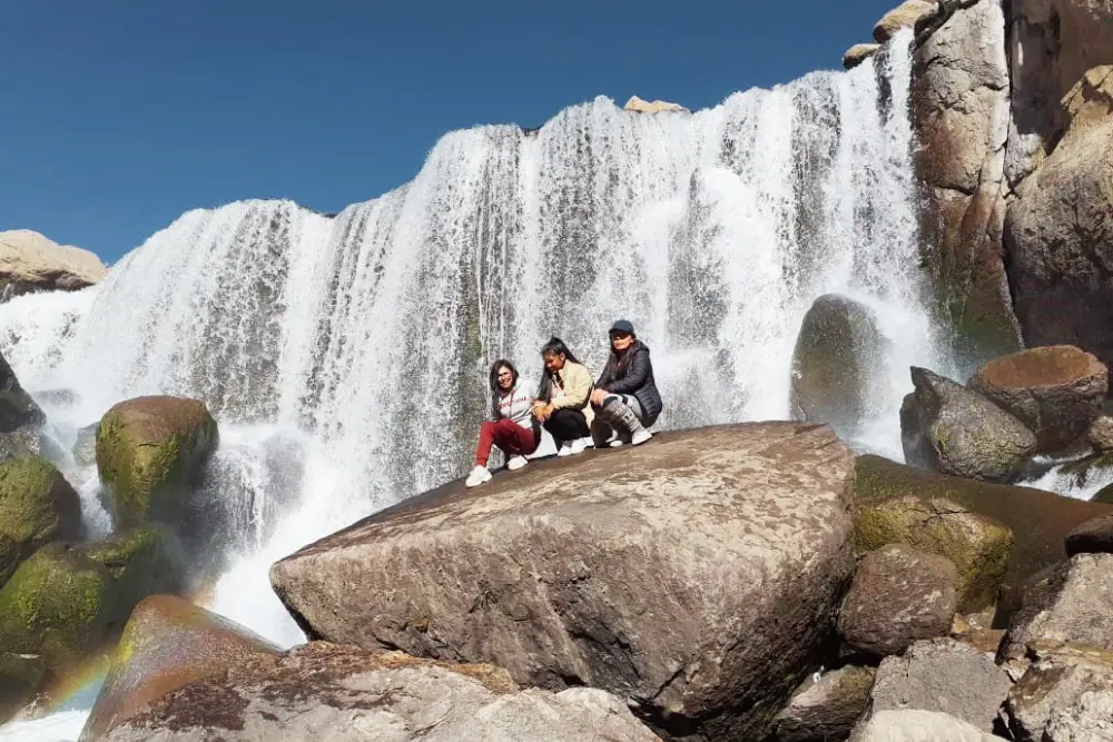Tres turistas sonrientes se toman una fotografía grupal frente a la caída de agua de las Cataratas de Pillones.